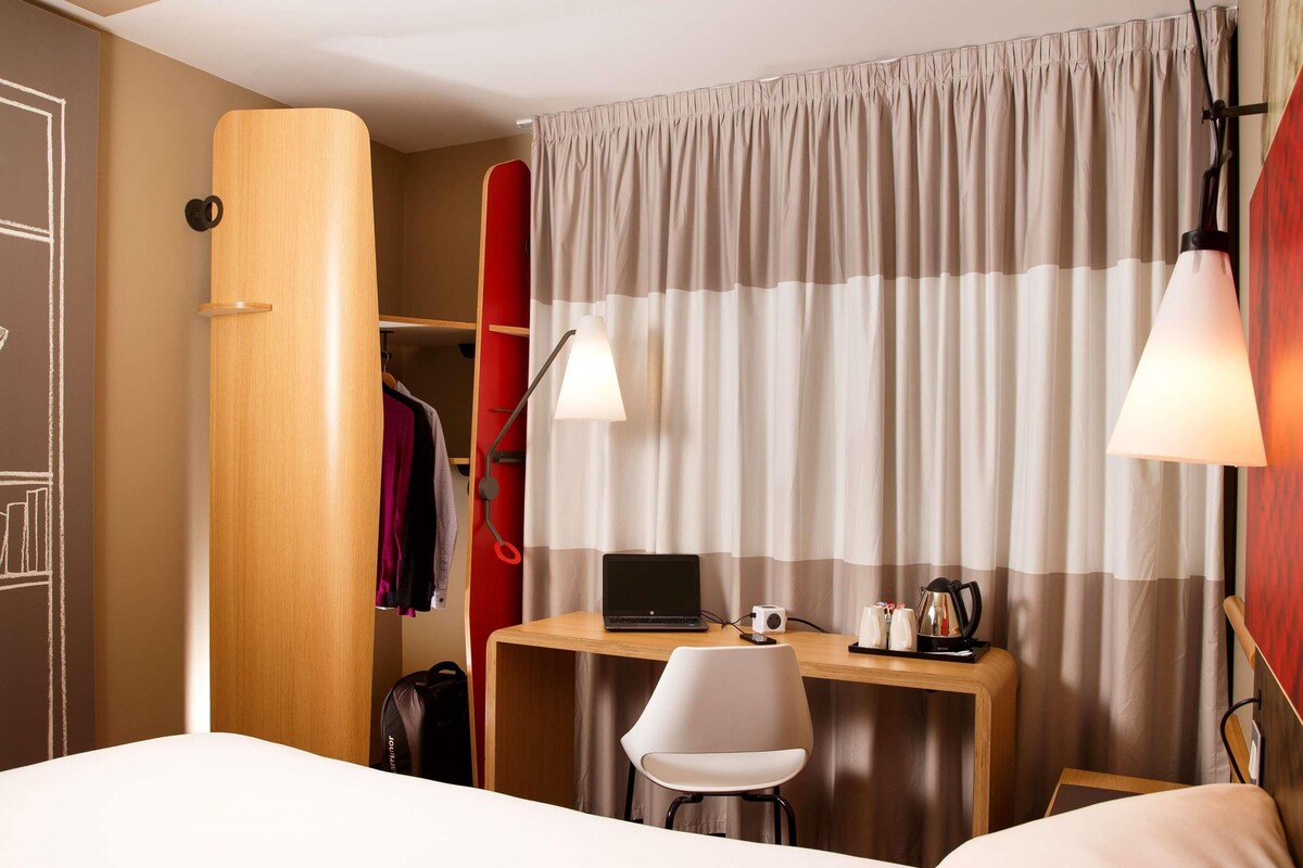 A hotel room with a bed, a desk with a laptop, a chair, a wardrobe, and a curtain.