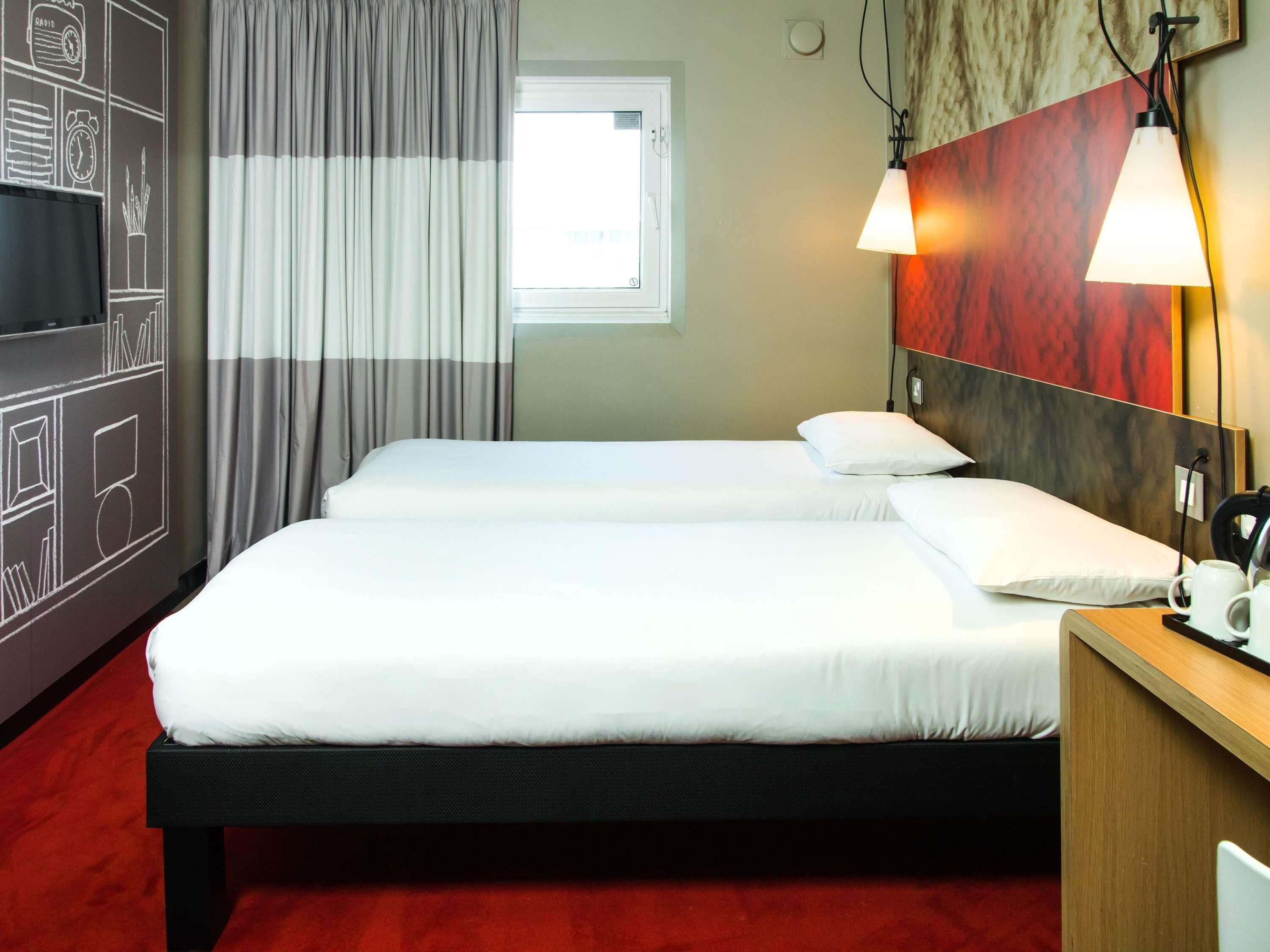 Een hotelkamer met twee bedden, een raam met gordijnen, een wandklok en een nachtkastje met een lamp.