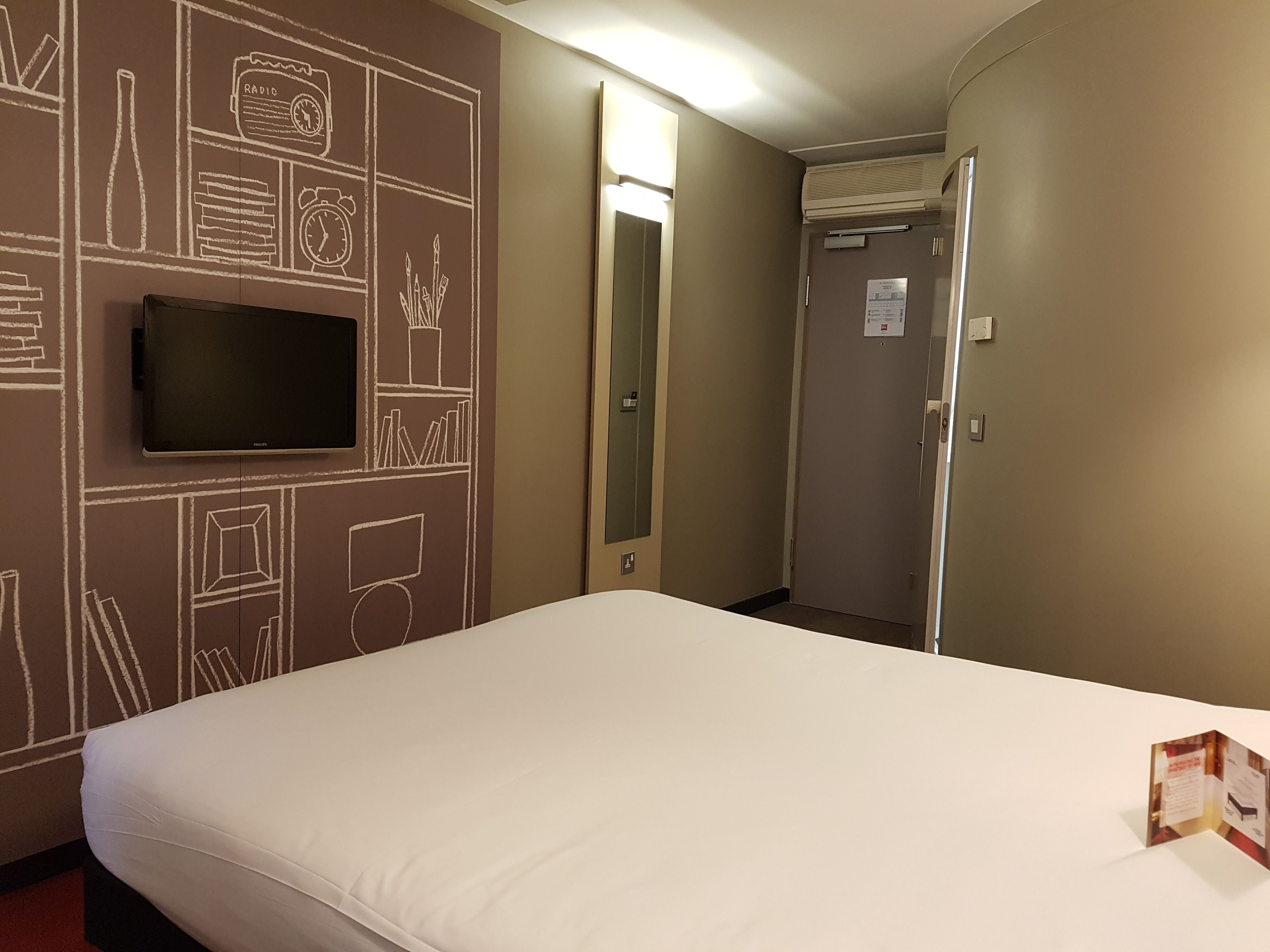 Habitación de hotel con una cama grande, un televisor de pantalla plana montado en la pared y una puerta que da a un pasillo.