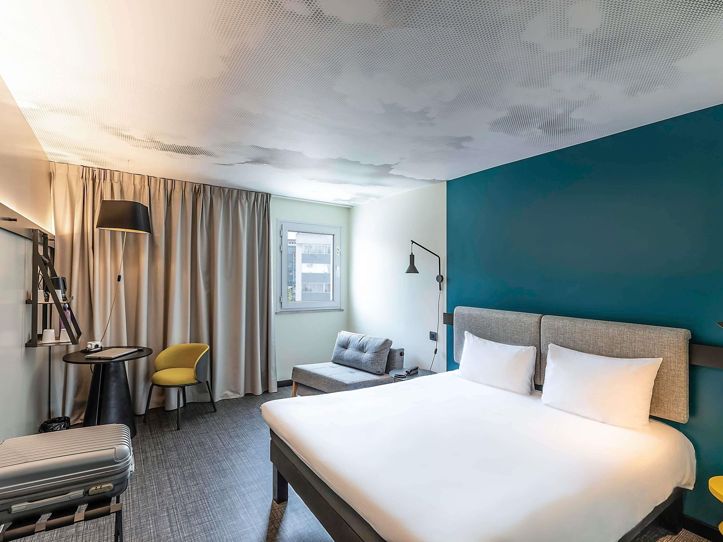 Quarto de hotel com cama, uma cadeira, escrivaninha, abajur e janela.