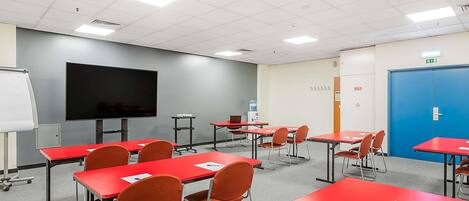 A classroom with red tables, chairs, a large screen, and a blue door.