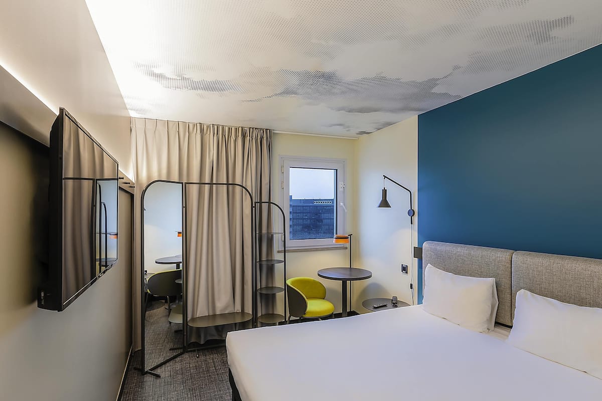 Une chambre d’hôtel avec un lit, un bureau, une chaise, une petite table, une fenêtre et un rideau.