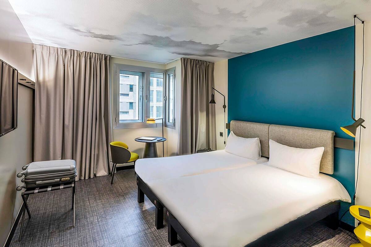 Een hotelkamer met een bed, een tafeltje, een stoel, een koffer en een raam met gordijnen.