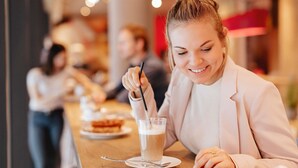 Una persona sentada a una mesa con una bebida y una cuchara, en un café con otras personas al fondo.
