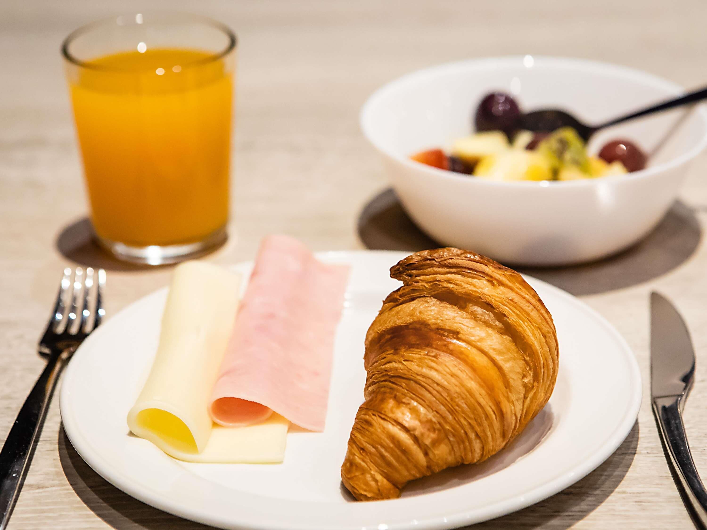 A plate with a croissant, ham, and cheese, a glass of orange juice, and a bowl of fruit.