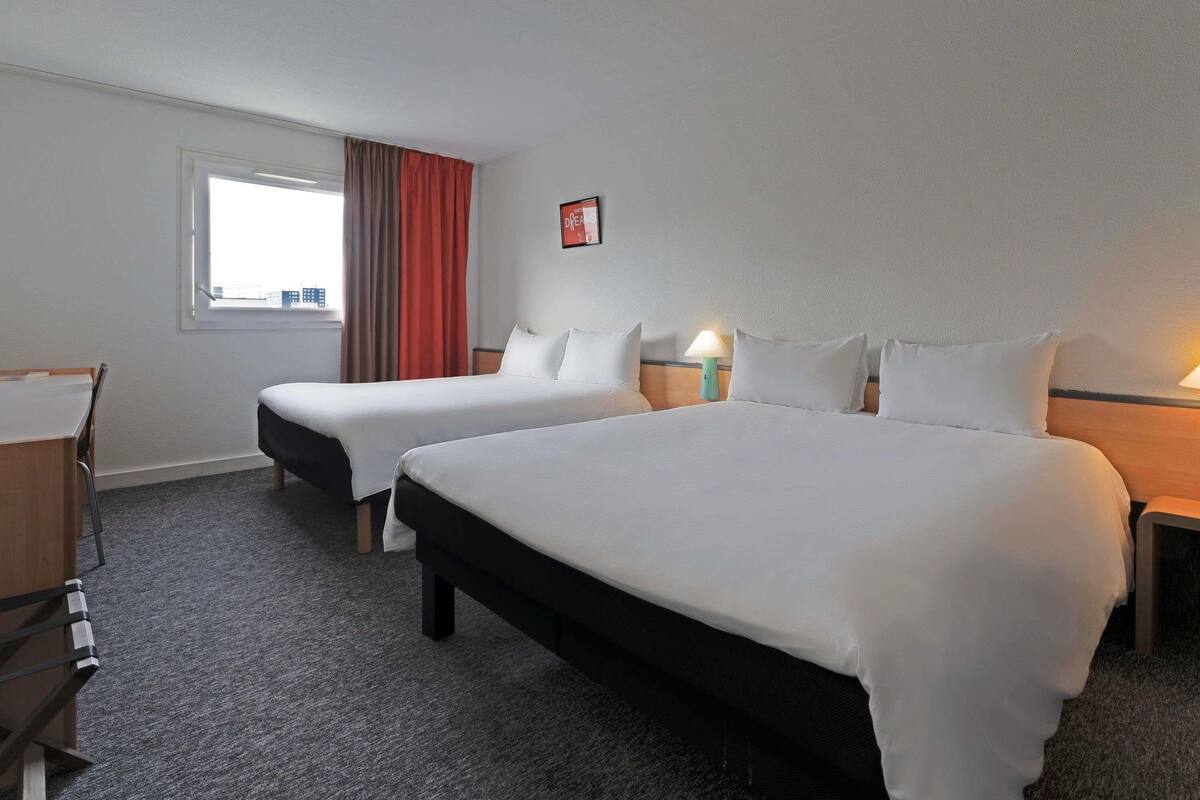 A hotel room with two beds, a desk, a chair, a window with curtains, and a wall-mounted picture.