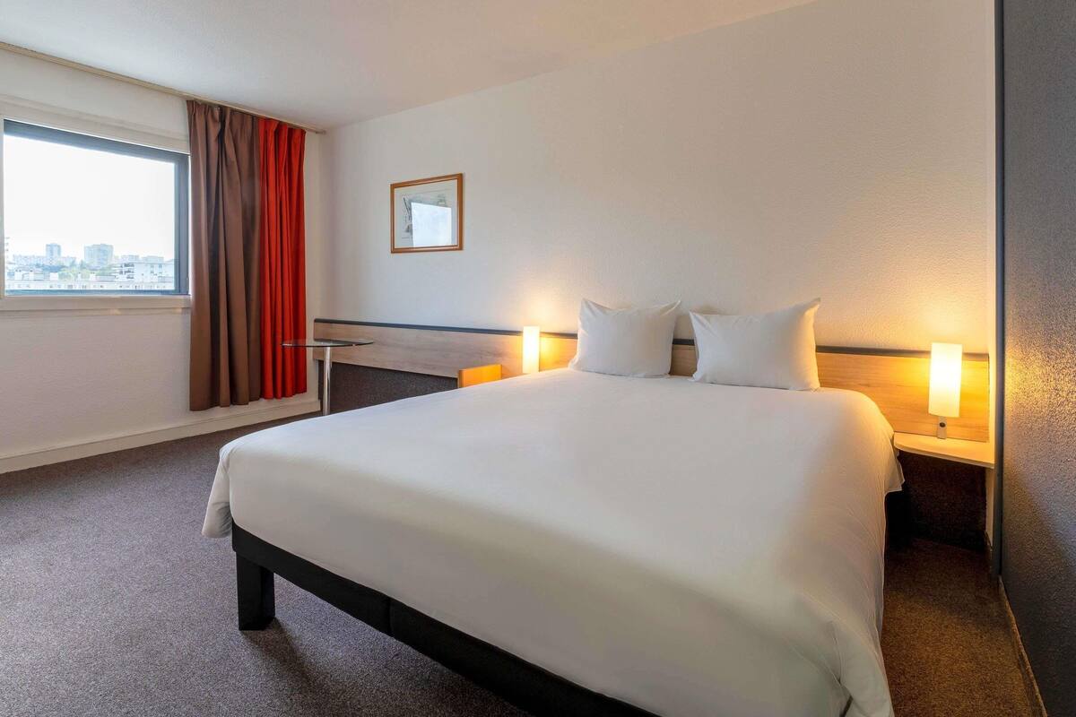 A hotel room with a large bed, two bedside lamps, a small table, and a window with curtains.