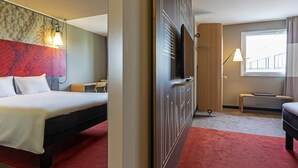 A hotel room with a bed, a desk, a chair, a TV, a window with curtains, and a red carpet. - ibis Paris Pantin Eglise (Pantin)