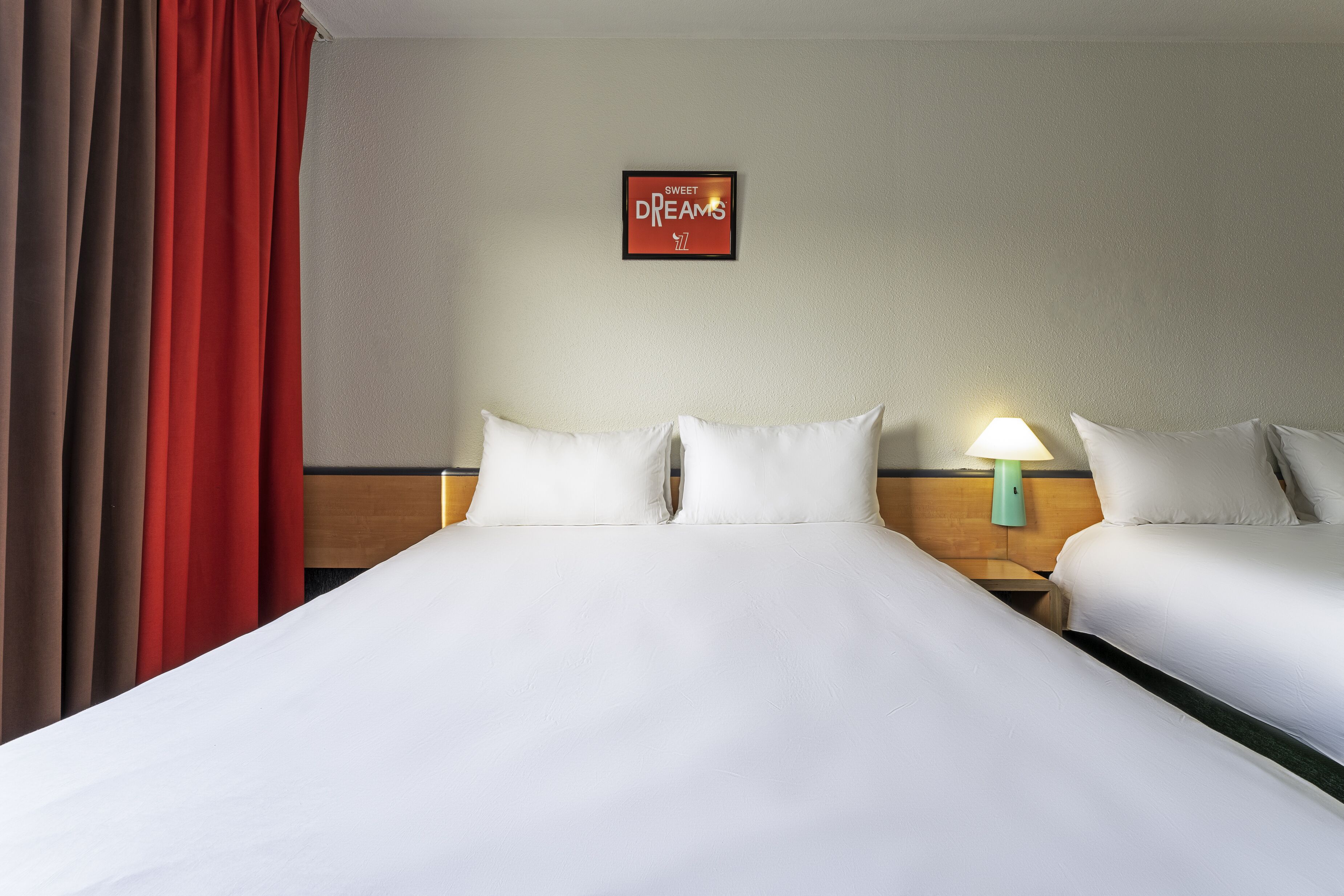 Habitación de hotel con dos camas, una cortina roja, un letrero en la pared y una mesita de noche con una lámpara.