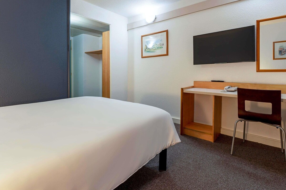 A hotel room with a bed, a desk, a chair, a flat-screen TV, and a framed picture on the wall.