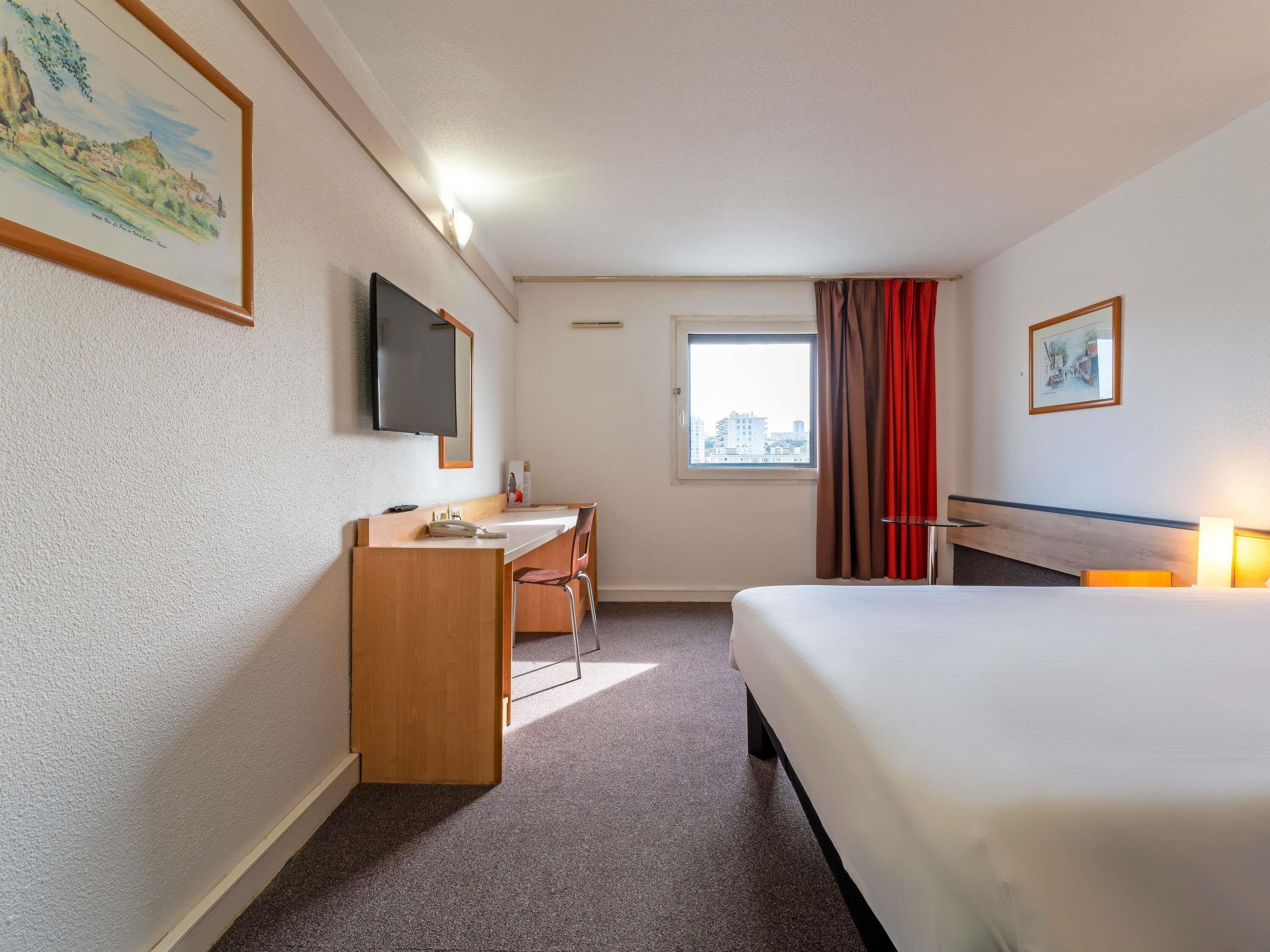A hotel room with a bed, a desk, a chair, a television, a window, and a painting on the wall.