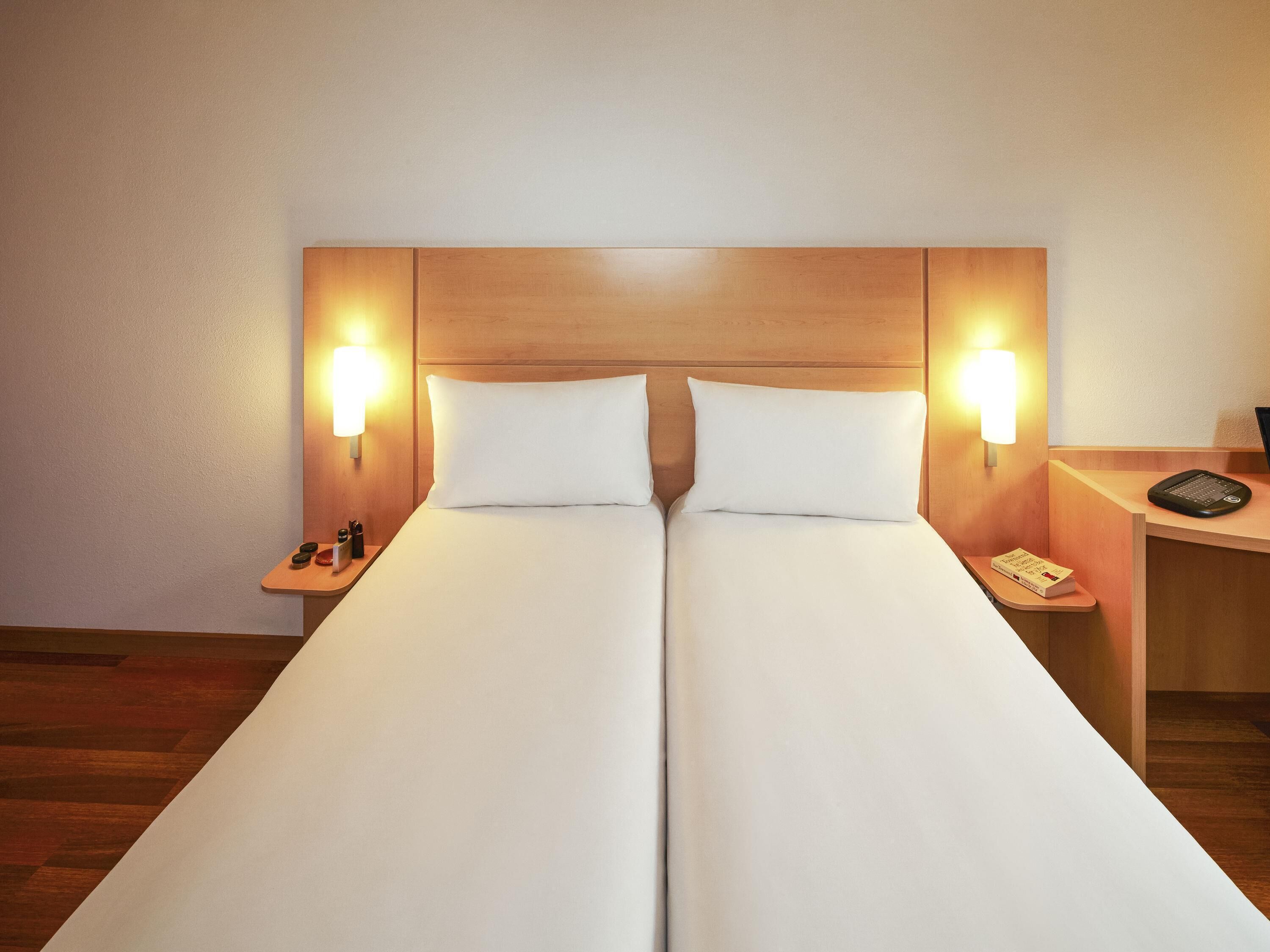 Una cama doble con dos almohadas, mesitas de noche con lámparas, una pequeña mesa con un libro y un teléfono, y un cabecero de madera.