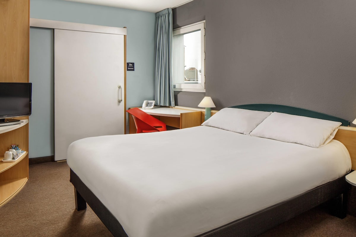A hotel room with a large bed, a desk with a chair, a television, and a shower sign.