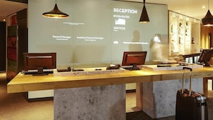 A reception desk with a digital screen displaying room rates and services, two computer monitors, and a suitcase on the floor. - ibis London Thurrock M25 (Grays)