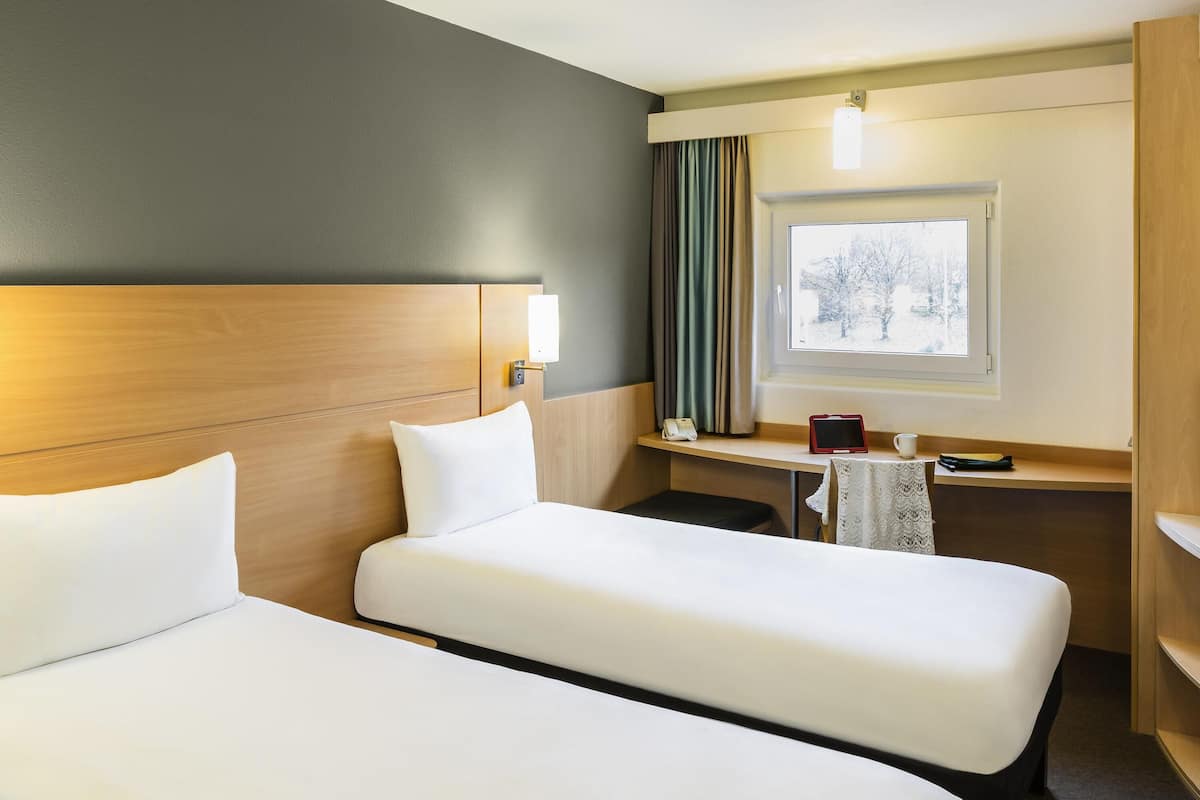 A hotel room with two beds, a desk, a chair, a window with a view, and a lamp.
