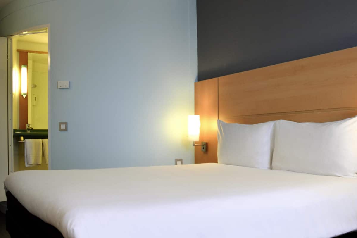A hotel room with a large bed, a nightstand, a wall-mounted lamp, and a bathroom visible through an open door.
