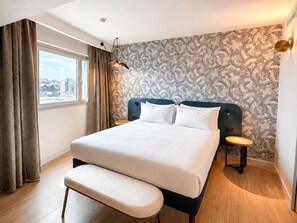 A modern hotel room with a large bed, a bench, a nightstand, a window with curtains, and a patterned wall. - ibis Antananarivo Ankorondrano (Antananarivo)