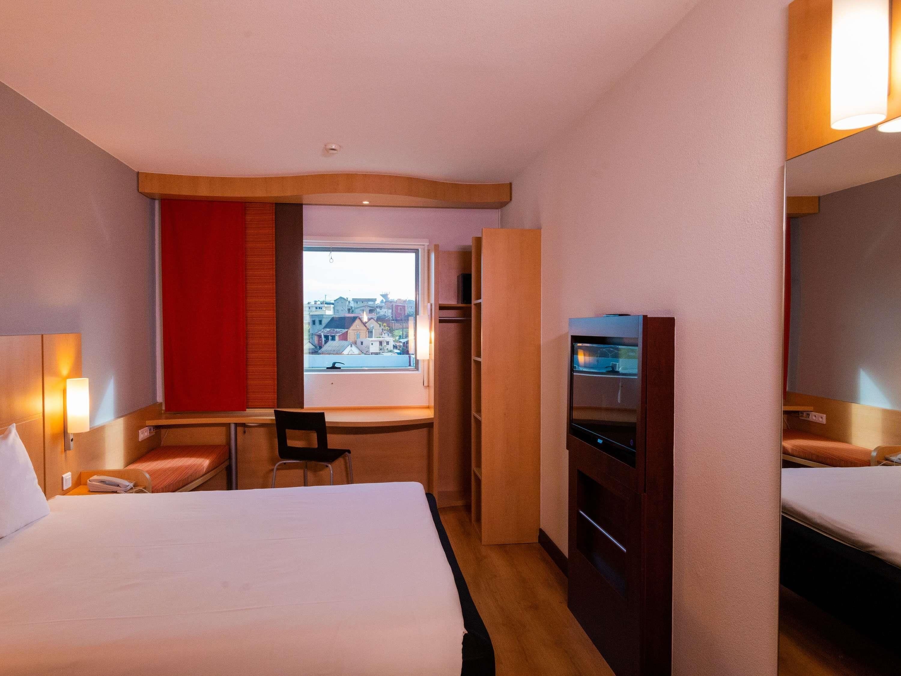 Une chambre d’hôtel équipée d’un lit, d’un bureau, d’une chaise, d’une télévision et d’une fenêtre donnant sur la ville.
