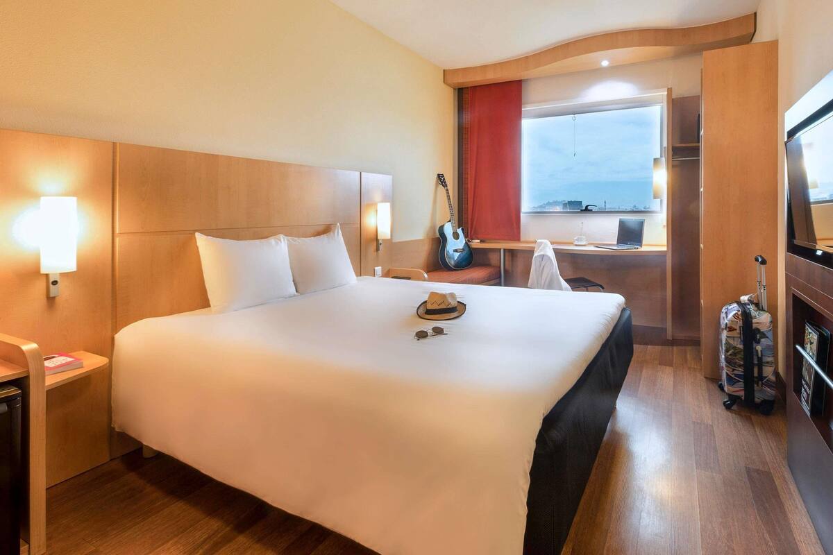 A hotel room with a large bed, bedside lamps, a desk with a laptop, a guitar, and a view of the sea.