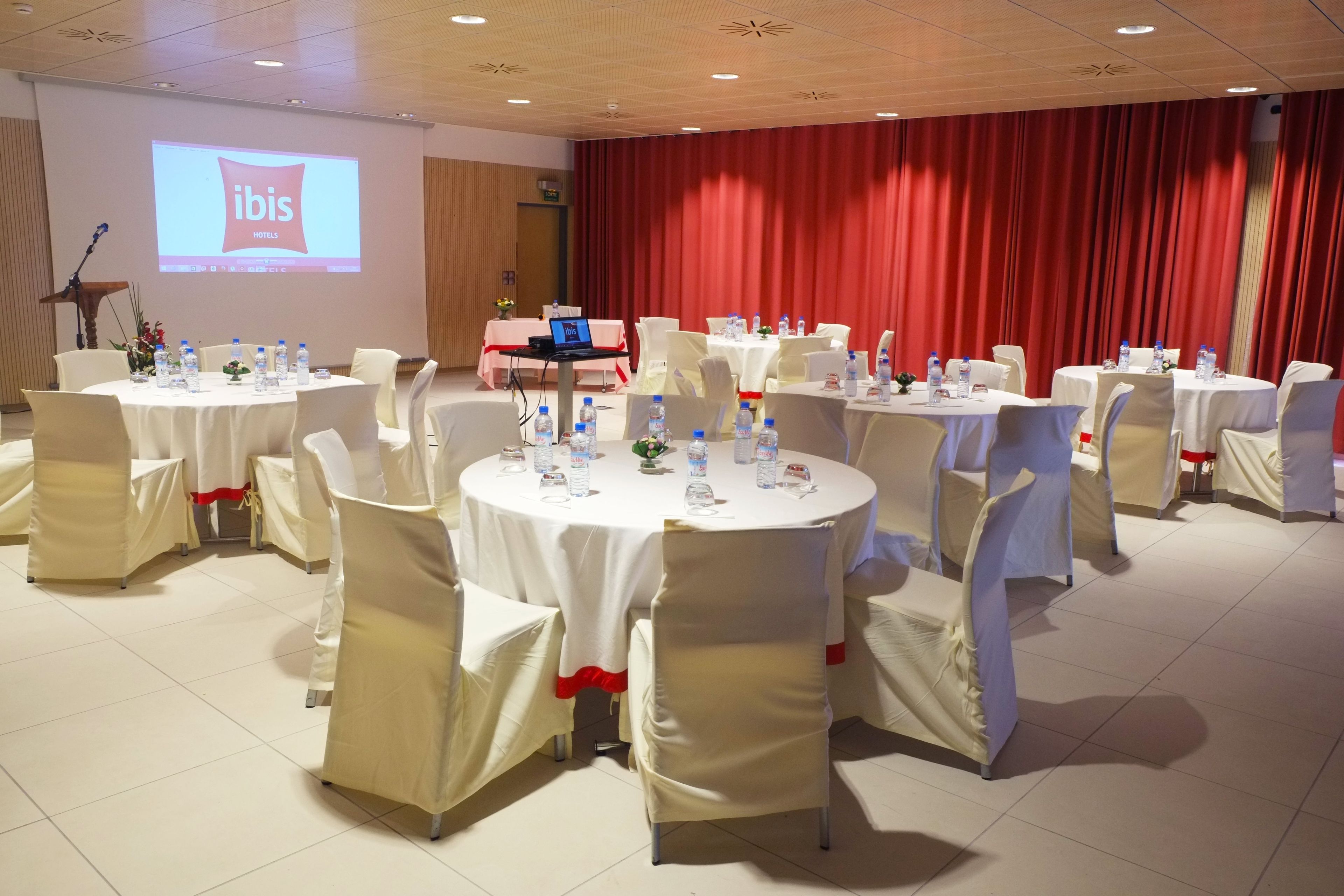 Un salón de conferencias con mesas redondas, sillas blancas y una pantalla grande que muestra 'ibis Hotels'.