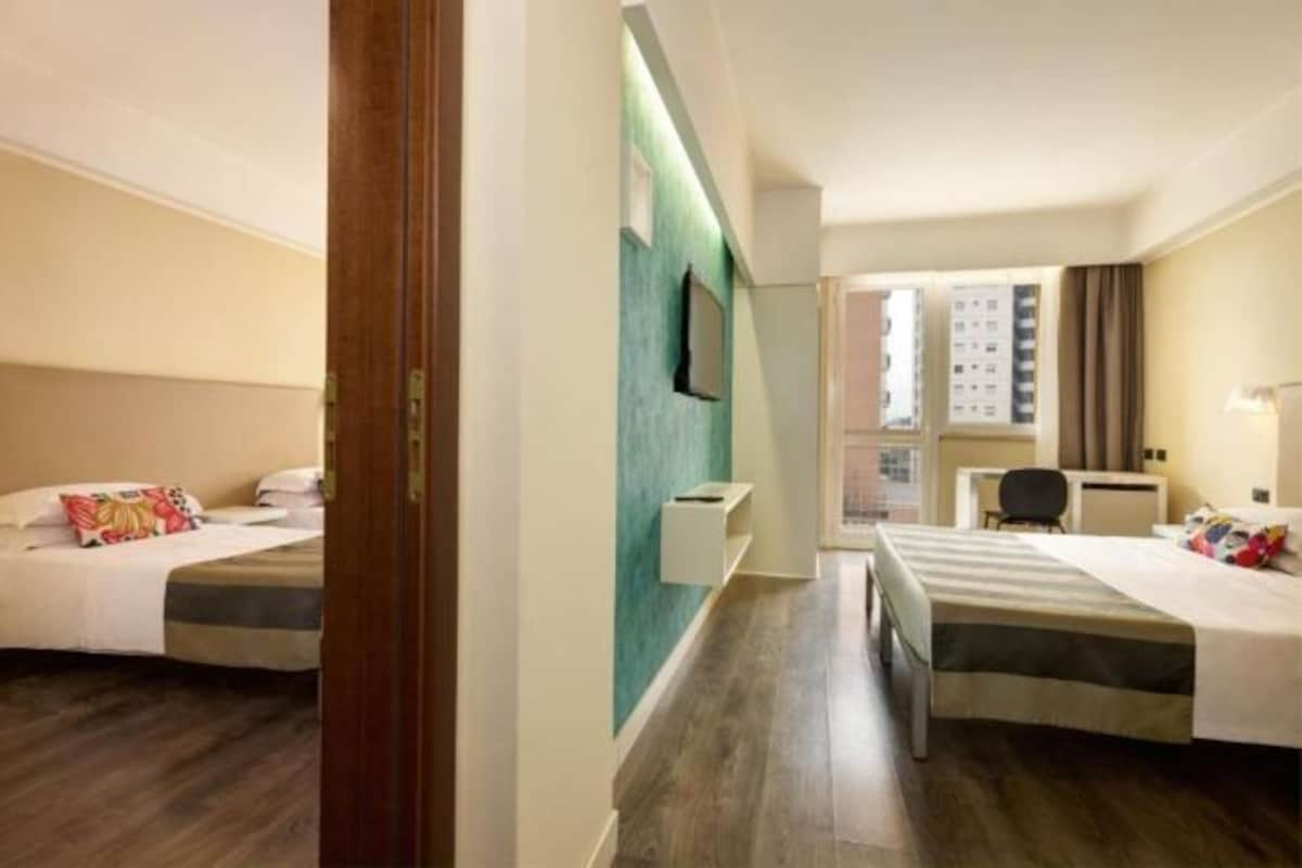 A hotel room with two beds, a desk, a chair, a TV, and a window.