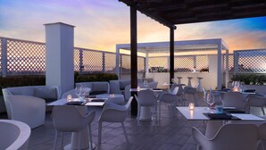 An outdoor dining area with white furniture and tables, set against a backdrop of a sunset sky. - Hotel Roma Tor Vergata (Rome)