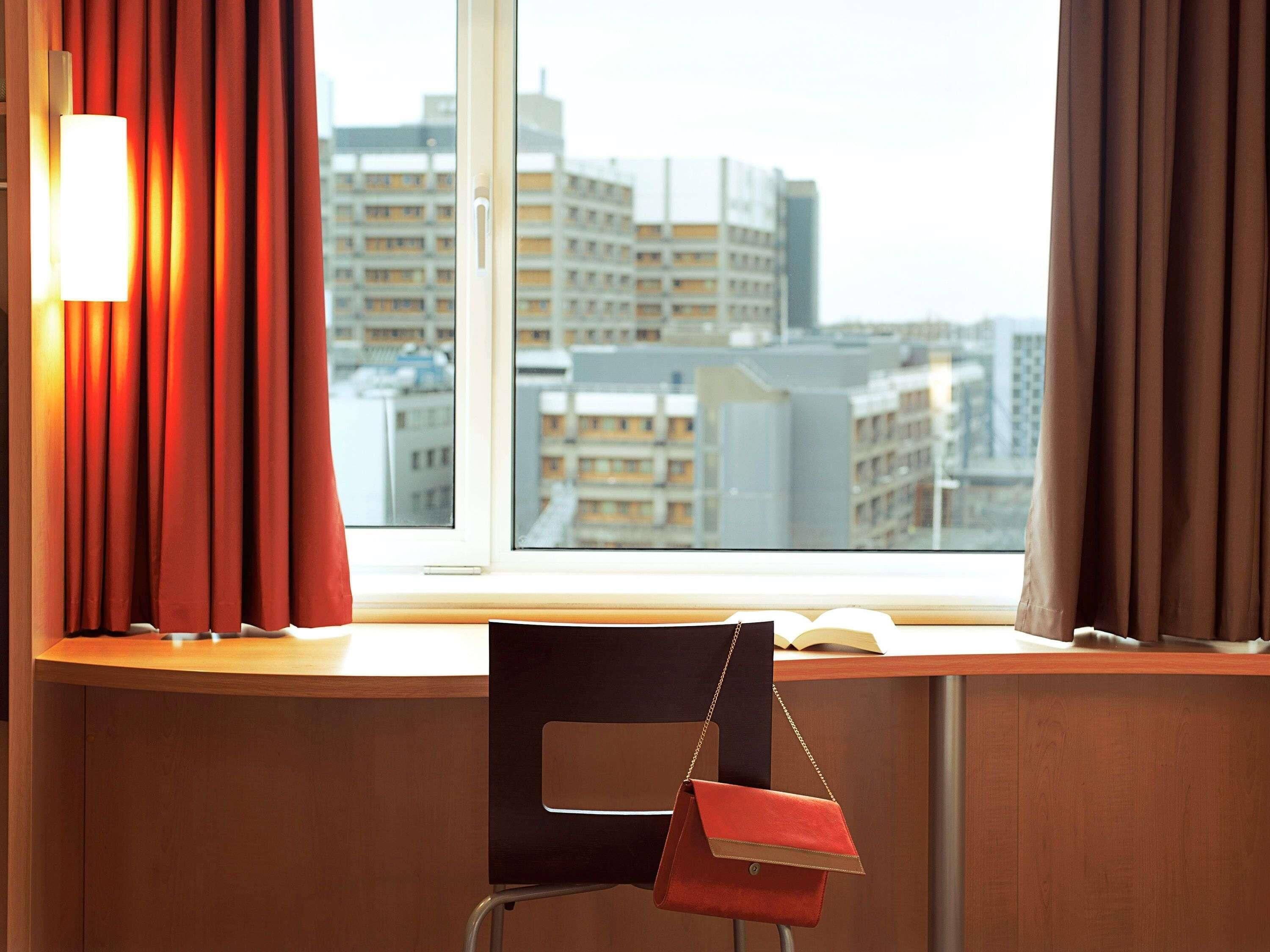 Hotellihuone, josta on näkymä kaupungin rakennuksiin, huoneessa on työpöytä ja tuoli sekä punainen käsilaukku, joka on tuolilla.