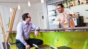 Zona de bar con un cantinero, un cliente sentado en un taburete y una pared verde con un diseño abstracto amarillo.