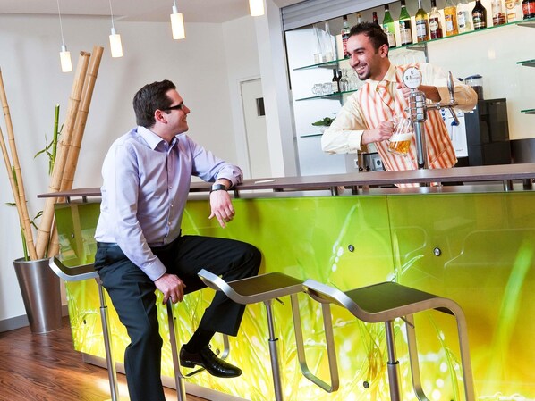 Zona de bar con un cantinero, un cliente sentado en un taburete y una pared verde con un diseño abstracto amarillo.