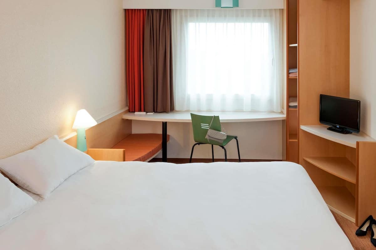 Una habitación de hotel con una cama, un escritorio con silla, televisión y ventana con cortinas.