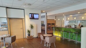 Un café moderno con suelo de madera, mesas redondas y taburetes de bar. Hay un televisor montado en la pared, plantas en macetas y una pequeña mesa con el menú.