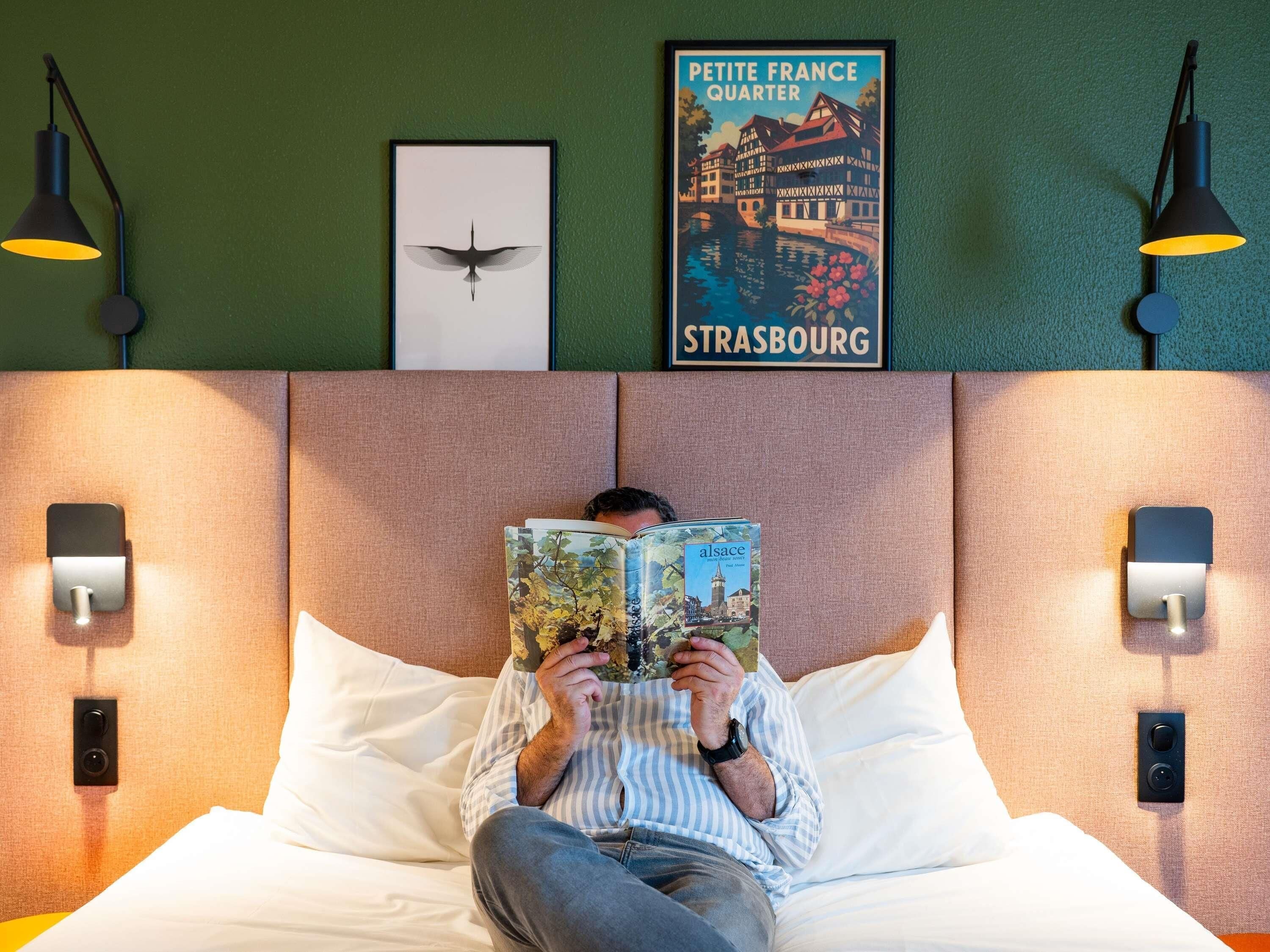 Uma pessoa lendo um livro num quarto de hotel com um quadro de Estrasburgo, uma cama e luminárias fixadas na parede.