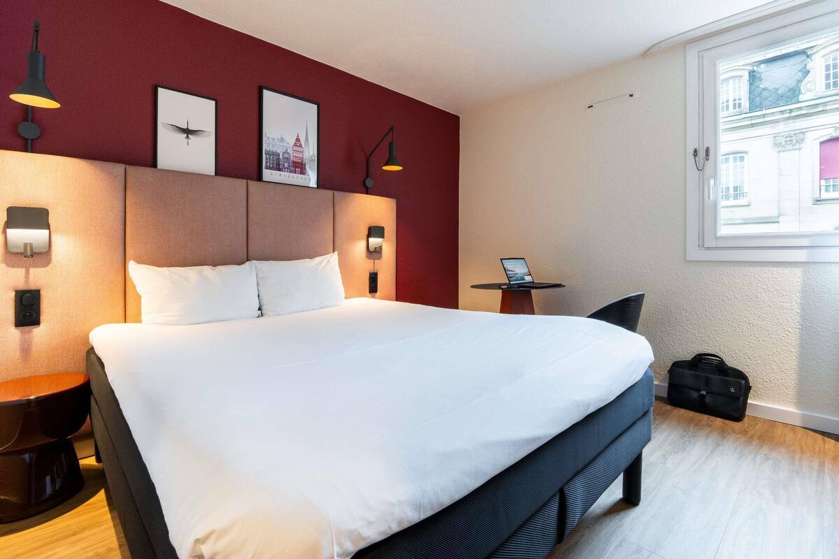 A hotel room with a large bed, a desk with a laptop, and a window with a view of buildings.
