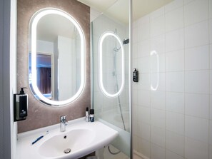 Un baño con un lavabo blanco, un espejo grande con luz y una ducha con puerta de cristal, además de una botella de champú en la pared.