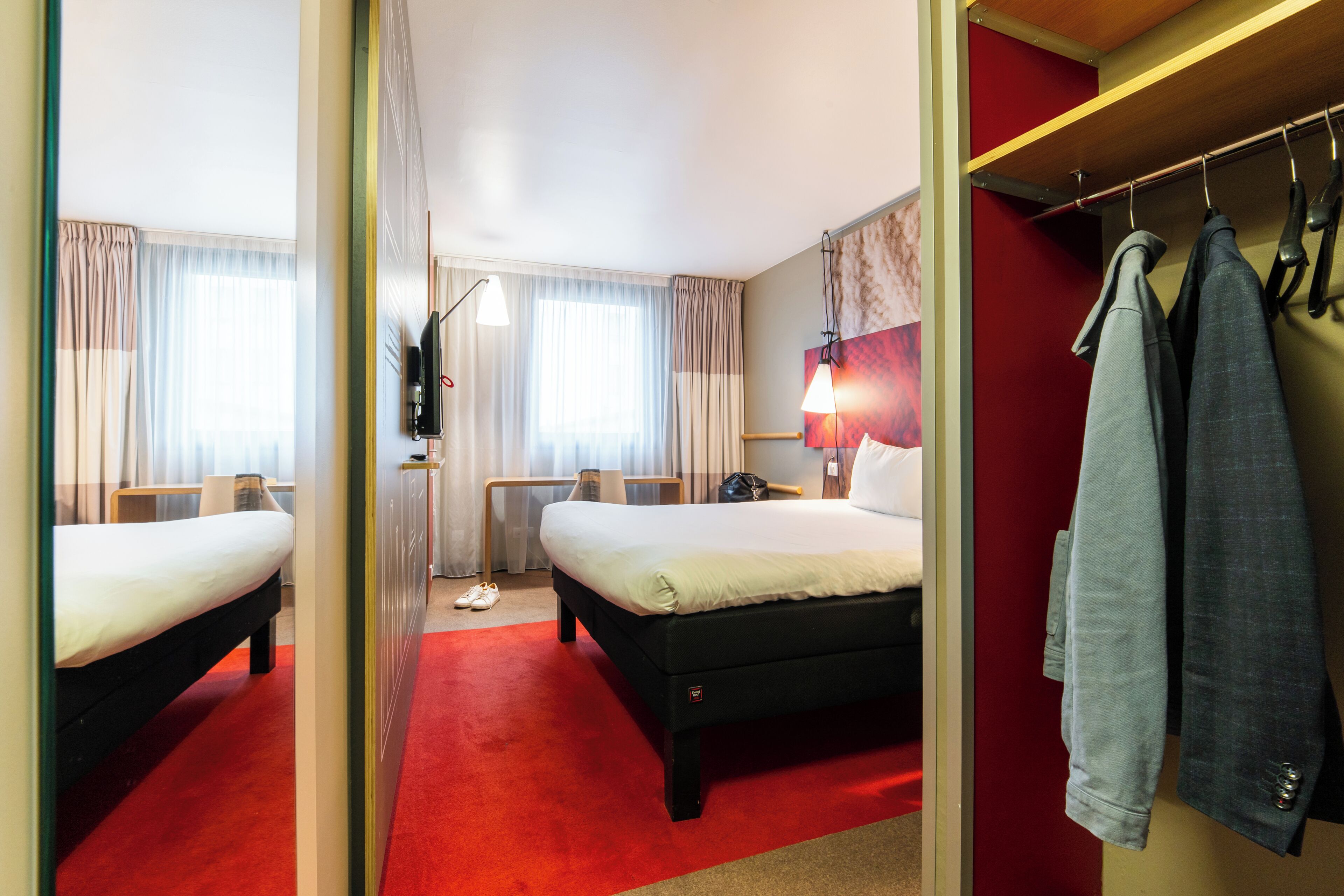 A hotel room with two beds, a red carpet, a mirror, a coat rack, and a television.