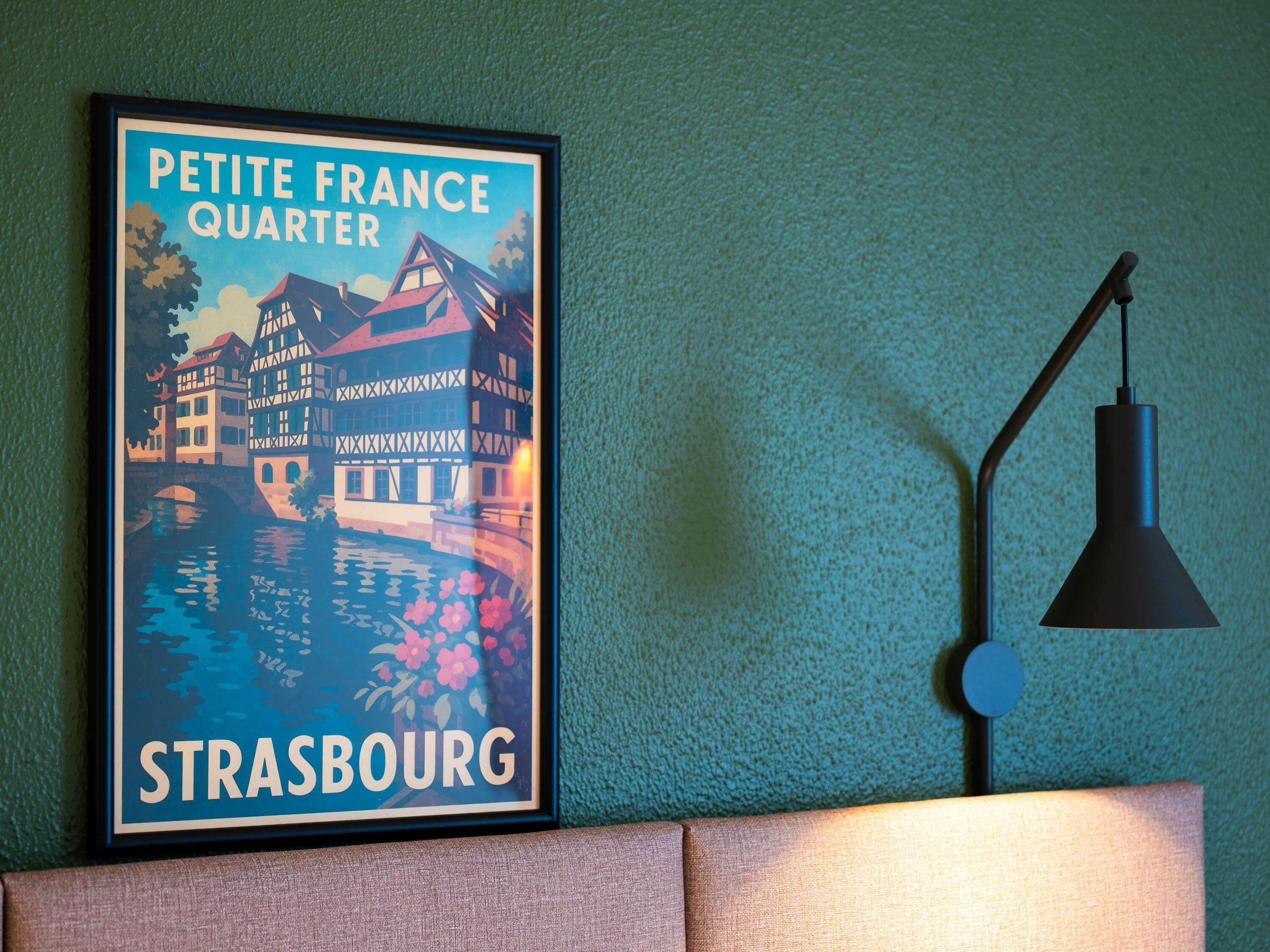 A framed poster of Strasbourg on a textured wall, a black desk lamp, and a cushioned surface.
