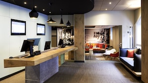 A modern reception area with a wooden reception desk, two computer monitors, and a seating area with a sofa and chairs. - Hôtel ibis Strasbourg Centre Petite France (Strasbourg)