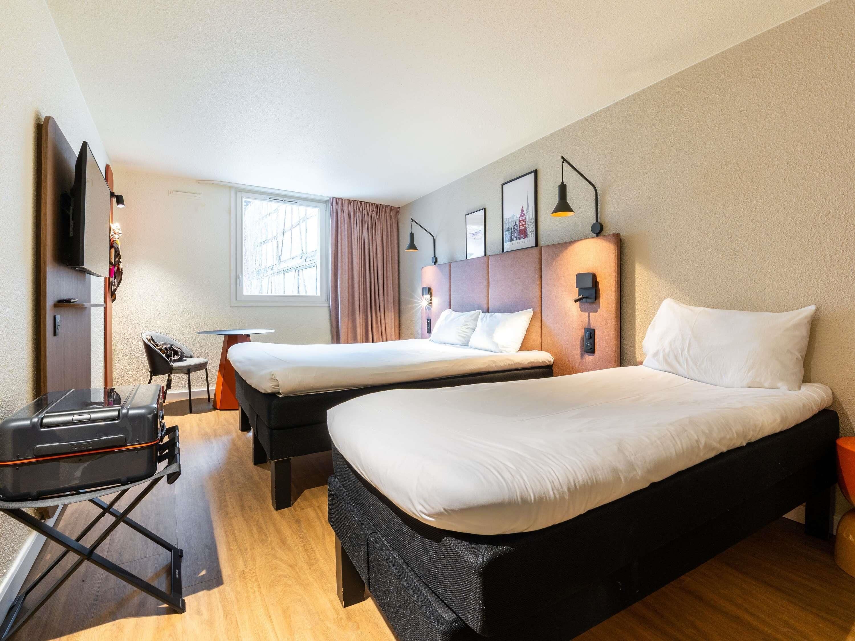 A hotel room with two beds, a desk, a chair, a TV, and a window with curtains.