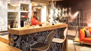 A modern bar area with a bar counter, hanging pendant lights, and a wall with abstract patterns. - Hôtel ibis Strasbourg Centre Petite France (Strasbourg)