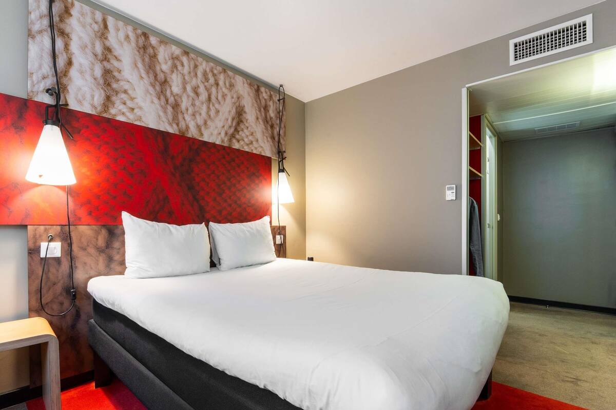 A hotel room with a large bed, two bedside lamps, a nightstand, a red carpet, and a red and beige textured wall.
