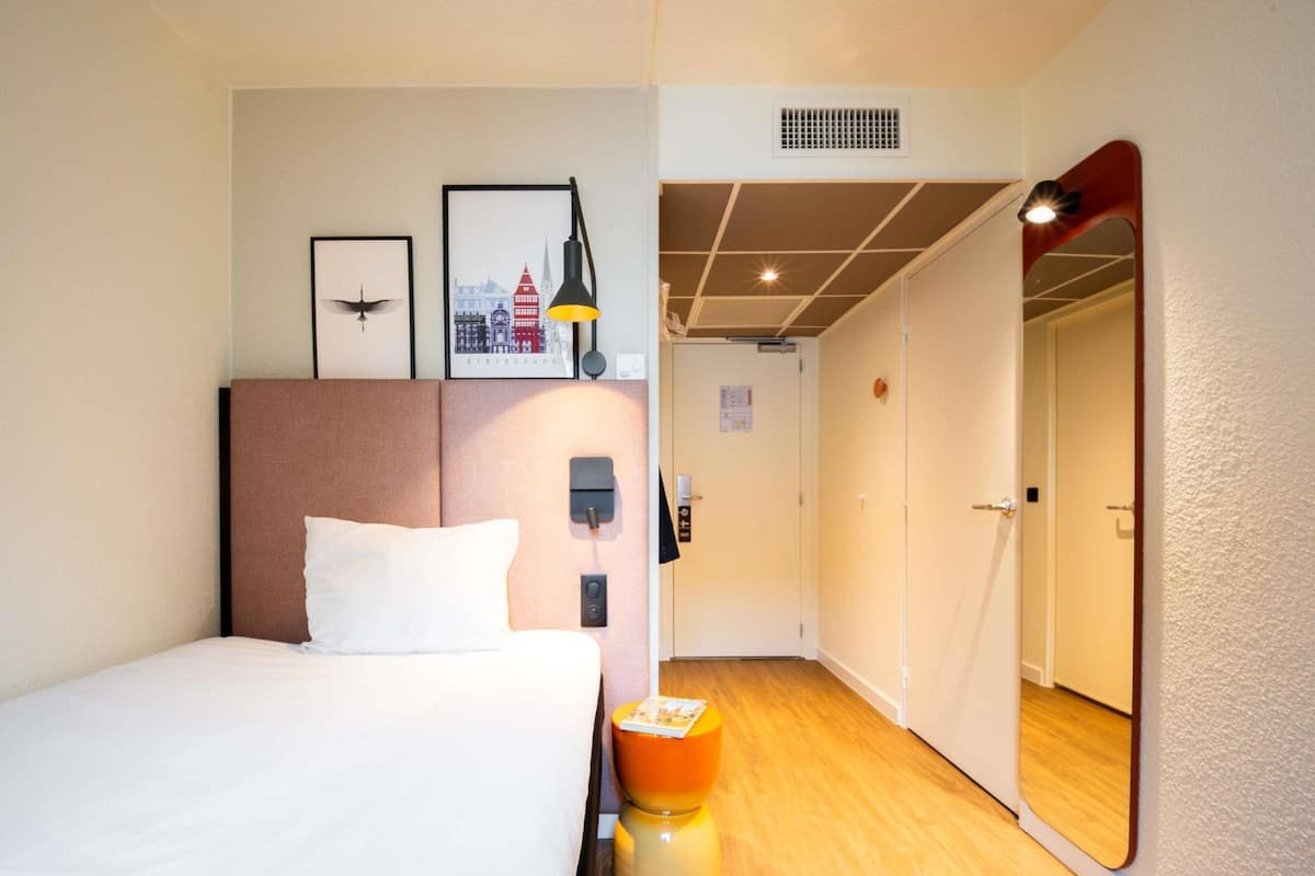 A hotel room with a bed, a nightstand, a mirror, a framed picture, a lamp, and a door leading to another room.