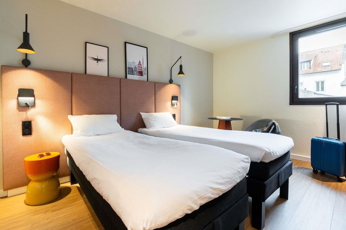 A hotel room with two beds, a small table, a chair, a suitcase, and a window with a view.