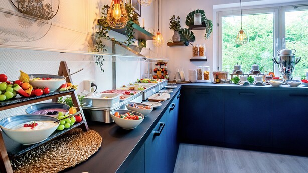 A modern kitchen with a breakfast buffet, wooden shelves, and a large window with a view of greenery. - DORMERO Hotel Weimar (Weimar)