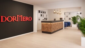 A modern reception area with a black wall displaying 'DORMERO' in red letters, a wooden reception desk, a blue sofa, and a potted plant. - DORMERO Hotel Weimar (Weimar)