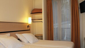A hotel room with a bed, a nightstand, a lamp, a window with curtains, and a television on the wall. - New Hôtel Saint Lazare (Paris)