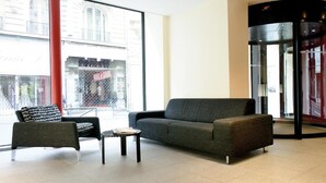 A modern lobby with a grey sofa, a black armchair, and a small table. A large window offers a view of a building with a sign that reads "BAIL & CEDER." - New Hôtel Saint Lazare (Paris)