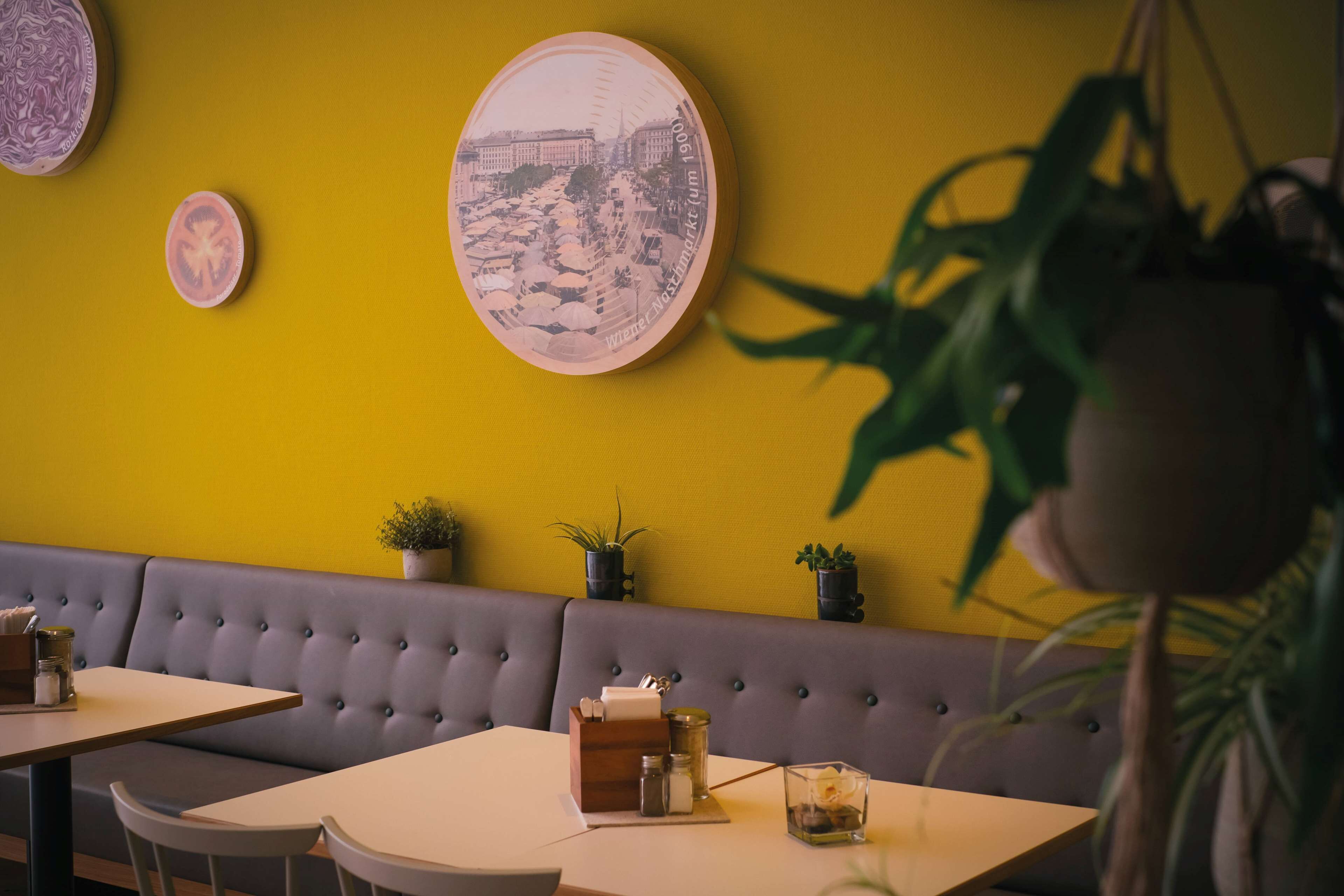 Interior de restaurante con pared amarilla, un cuadro redondo grande y un banco tapizado en gris.