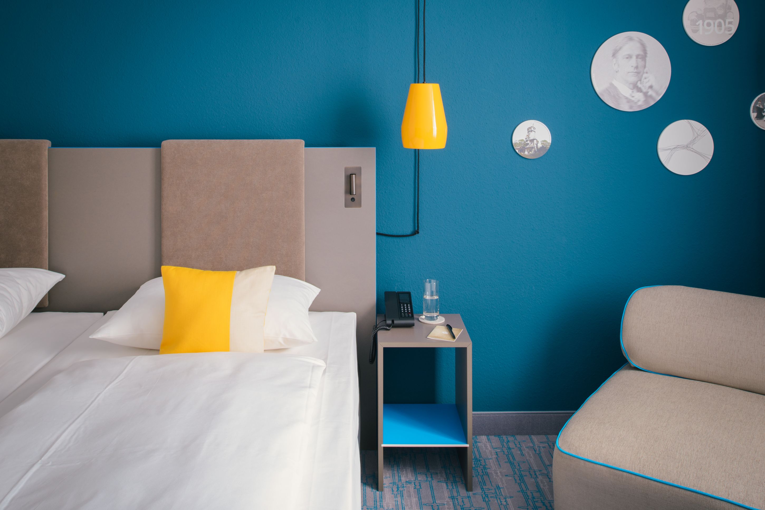 Une chambre d’hôtel avec un lit, une table de chevet avec un téléphone et un verre, une chaise, et un mur bleu orné de motifs circulaires.