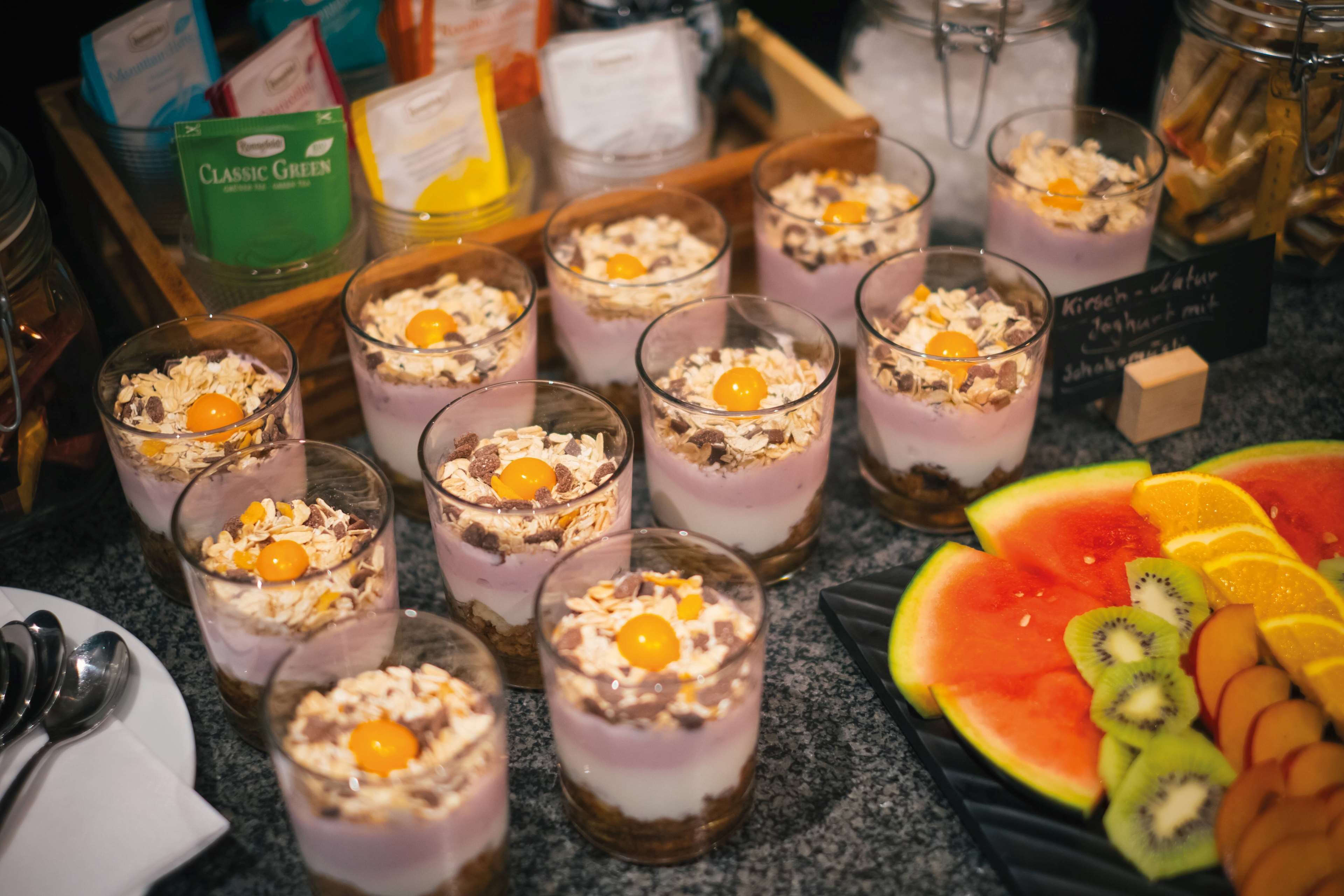 Diversos alimentos, incluyendo postres en capas en vasos, frutas frescas, y un letrero con texto en un idioma extranjero.