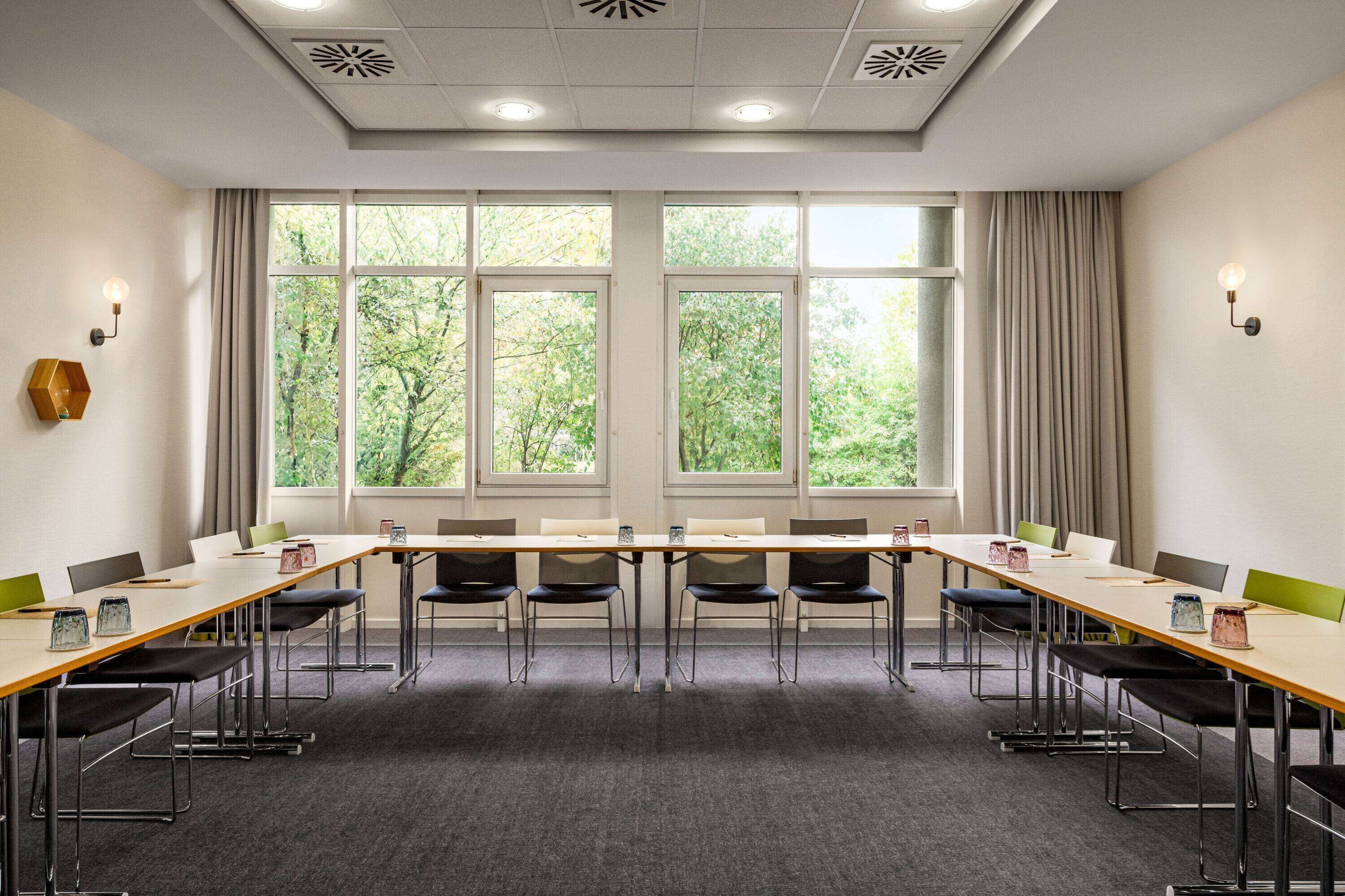 Un salón de conferencias con una larga mesa, sillas y ventanas grandes que dan a un paisaje de árboles.